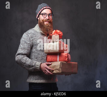 Portrait of bearded male in eyeglasses dressed in a warm wool sweater and a hat holds Christmas gifts over grey background. Stock Photo