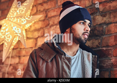 Portrait of bearded Indian hipster male dressed in a warm jacket and a hat, drinks coffee from a red cup near the wall of a red brick. Stock Photo