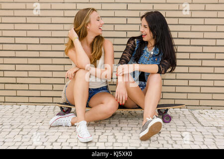 Two beautiful and young girlfriends having fun with a skateboard, in front of a brick wall Stock Photo