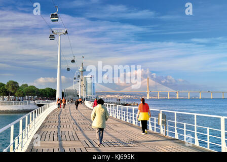 People passing pedestrian bridge surrounded by modern architecture, cable car lift and Vasco da Gama bridge Stock Photo