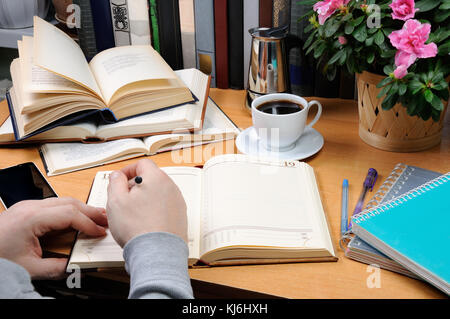 a man holds a pen and takes notes in a notebook on a desk in his office, surrounded by books, notebooks with a cup of black coffee Stock Photo