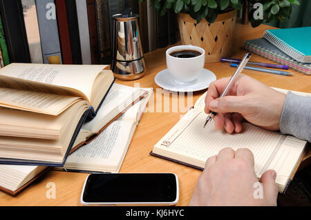 a man holds a pen and takes notes in a notebook on a desk in his office, surrounded by books, notebooks with a cup of black coffee Stock Photo