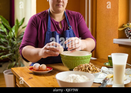 Close up photo of young female hands breaking an egg by the edge of the green bowl, bowl full of wallnuts is placed close by and  ingredients for baki Stock Photo