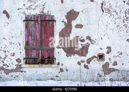 An old window with closed red worn wooden shutters on a white decrepit wall in winter with copy space. Stock Photo