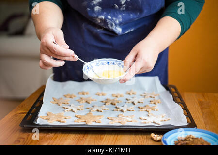 Woman baking at home ginger bread cookies in a shape of star for Christmas.Focus on hands holding a small bowl, mixing an egg with a fork, tray full o Stock Photo