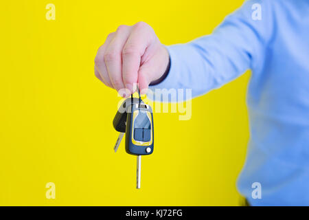 close up photo of male hand holding a car key. Person is wearing a blue shirt, standing in front of the bright yellow background Stock Photo