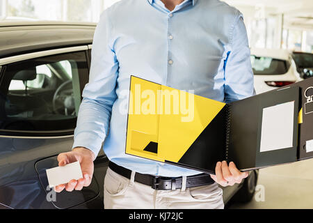 Close up of male adult car dealer standing indoors at car dealership,holding an opened blank booklet and business card in his hands, cars at the backg Stock Photo