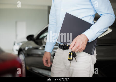 Close up of male adult car dealer standing indoorts at car dealership,holding a folder and car keys in his hand with a new car behind him Stock Photo