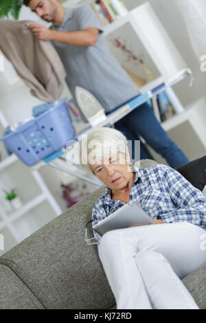 Shocked woman looking at tablet, man ironing in background Stock Photo