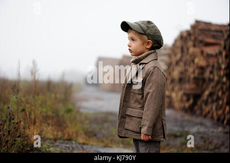 A sad,pretty, four years old boy lumberjack playing  on pill of wood background. Foggy gloomy day. Work, working, job, transport boy is  looking ahead Stock Photo