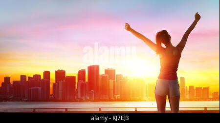 Jogging, Fitness And Success - Girl With Arms Raised And Cityscape At Sunset Stock Photo