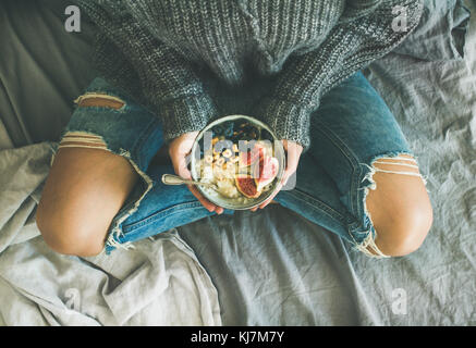 Healthy winter breakfast in bed. Woman in sweater and jeans holding rice coconut porridge with figs, berries, hazelnuts, top view. Clean eating, veget Stock Photo