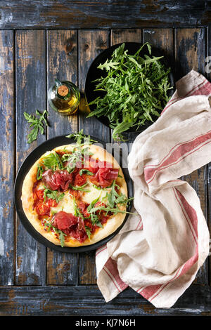 Whole homemade pizza with cheese and bresaola, served on black plate with fresh arugula, olive oil and kitchen towel over old wooden plank background. Stock Photo