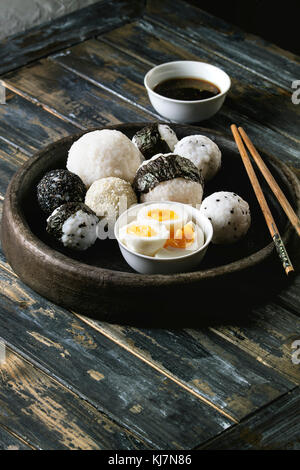 Stone tray with different size rice balls with black sesame and seaweed nori, served with soft boiled eggs, soy sauce, chopsticks over wooden plank ba Stock Photo