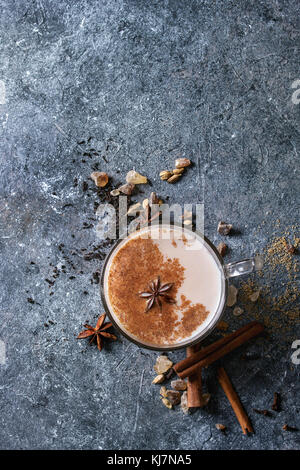 Glass cup of traditional indian masala chai tea with ingredients above. Cinnamon, cardamom, anise, sugar, black tea over dark texture background. Top Stock Photo