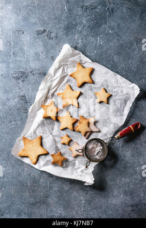 Homemade shortbread star shape sugar cookies different size with sugar powder on baking paper with vintage sieve over blue texture Background. Christm Stock Photo