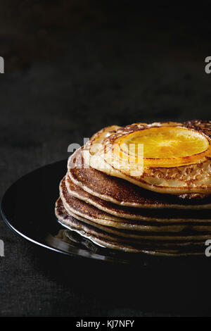 Stack of homemade pancakes with fried orange, served with mapple syrup on black plate over dark background. Close up Stock Photo