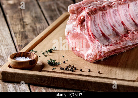 Fresh raw uncooked whole rack of pork loin with ribs on wooden cutting board with salt, thyme and butcher clever over old wood plank table. Close up. Stock Photo