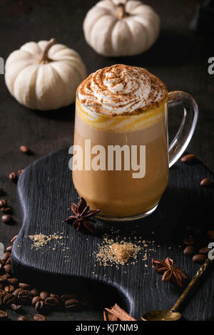 Glass of spicy pumpkin latte with whipped cream and cinnamon standing on black serving board with decorative white pumpkins. Coffee beans and spices a Stock Photo