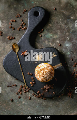 Glass of spicy pumpkin latte with whipped cream and cinnamon standing on black serving board. Coffee beans and spices above. Dark background. Top view Stock Photo