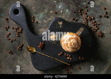 Glass of spicy pumpkin latte with whipped cream and cinnamon standing on black serving board. Coffee beans and spices above. Dark background. Top view Stock Photo