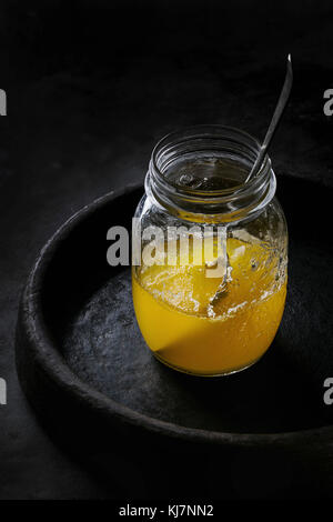 Glass of hot drawn ghee butter standing with spoon in clay terracotta tray over dark background. Healthy eating Stock Photo