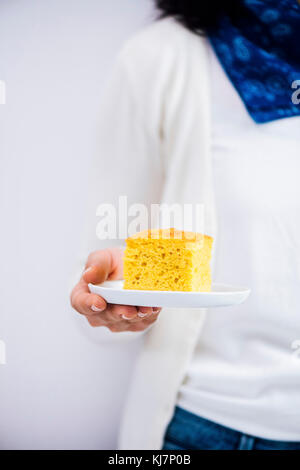 A woman in white shirt and white sweater with a blue scarf holding a slice of pumpkin cornbread on a white plate photographed from front view. Stock Photo