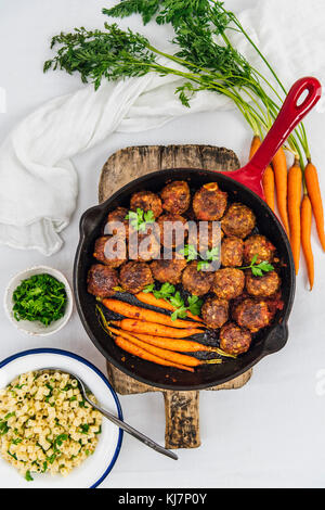 Gluten-Free Honey Garlic Meatballs and roasted baby carrots in a cast iron pan placed on a wooden cutting board photographed from top view. Raw baby c Stock Photo