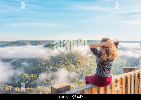 Back of young woman sitting on railing by mountains and floating fog clouds in morning in Grandview Overlook, West Virginia during golden autumn with  Stock Photo