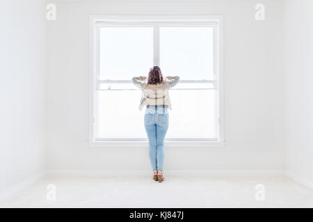 Back of one young woman standing in white, bright empty, clean room on carpet of modern new house, home, apartment looking through the sunny large win Stock Photo