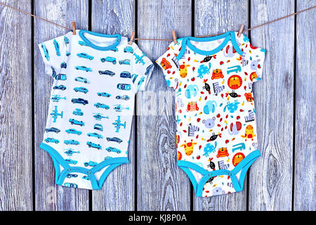 Infants cute rompers hanging on rope. Baby patterned bodysuits drying on clothesline on old wooden background. Stock Photo