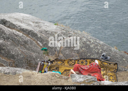 Looking down at partial view of 2 unrecognizable people seated on a colorful patterned blanket on a rock enjoying view of the water below. Stock Photo