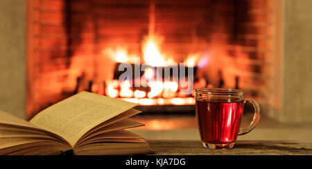 Relaxation at home. Close up of a cup of tea and a book on a burning fireplace background Stock Photo