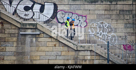 Bremen, Germany - March 8th, 2017 - Two construction workers wearing hi-viz vests and helmets walking down a set of stairs in front of an old graffiti Stock Photo