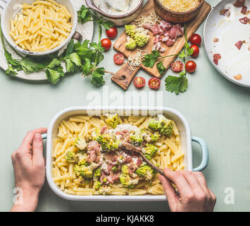 Woman female hands with spoon cooking pasta casserole with romanesco cabbage and ham in creamy sauce on kitchen table background with ingredients, top Stock Photo