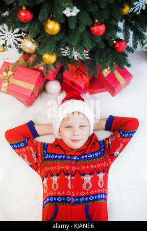 Portrait of cute funny little kid dressed in red pajamas and hat of santa. White happy kid laying on white wooden floor near holiday presents and tree Stock Photo