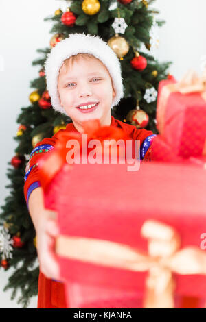 Closeup portrait of happy smiling child holding pile of present boxes in hands. Boy wearing santa hat and red pajamas. Holiday green tree in backgroun Stock Photo