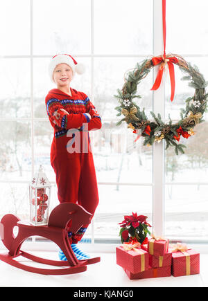 Portrait of cute little child in holiday christmas white and red interior. Happy smiling kid posing for photo standing near big window with snowy city Stock Photo