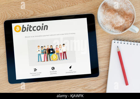 The Bitcoin website features on an iPad tablet device which rests on a wooden table beside a notepad and pencil and a cup of coffee (Editorial only) Stock Photo