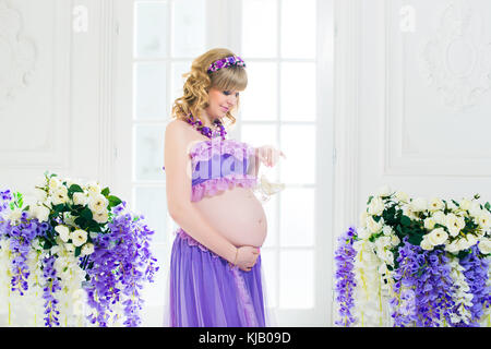 A happy pregnant woman in a purple long skirt poses in the studio against a background of flowers. Stock Photo