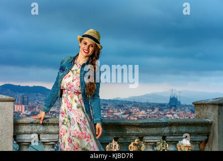 Perfect evening with stunning view. Portrait of happy elegant traveller woman in long dress and straw hat against cityscape of Barcelona, Spain Stock Photo
