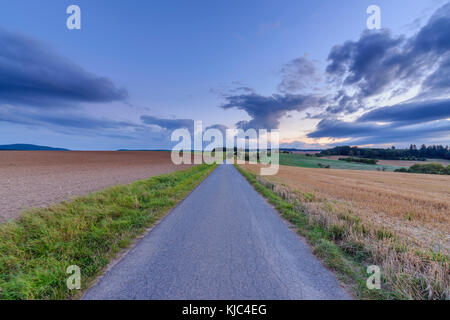 Countryside with harvested cereal field and paved laneway at dusk in summer at Roellbach in Spessart hills in Bavaria, Germany Stock Photo