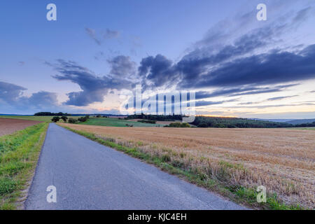 Countryside with harvested cereal field and paved laneway at dusk in summer at Roellbach in Spessart hills in Bavaria, Germany Stock Photo