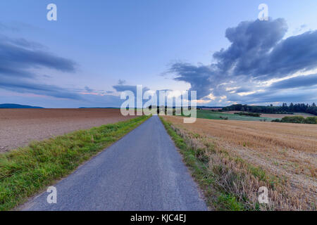 Countryside with harvested cereal field and paved laneway at dusk in summer at Roellbach in Spessart hills in Bavaria, Germany Stock Photo