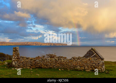 Remains of a stone house on the Isle of Skye with a rainbow appearing over the coast in Scotland, United Kingdom Stock Photo