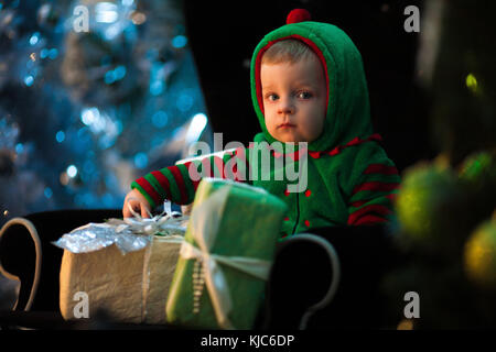 Little boy in green hood sits in black armchair with Christmas gift boxes on background of celebration lights. Stock Photo