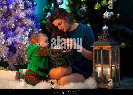 Happy mother with her little son sit on white fur rug and look at Christmas gifts in wicker basket on background of Christmas tree, decorations and li Stock Photo