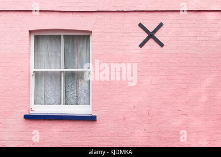 Victorian window with white wooden sash, white curtains and blue balcony on a bright pink painted brick wall with metal black cross on it. Stock Photo