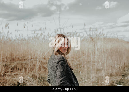 Pretty and young woman with long blonde hair dressed in wool coat looking at camera over her shoulder and smiling. Background bulrush. Autumn. Outdoor Stock Photo