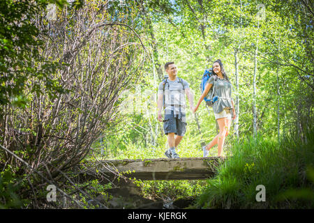 Couple carrying backpacks across bridge in woods Stock Photo